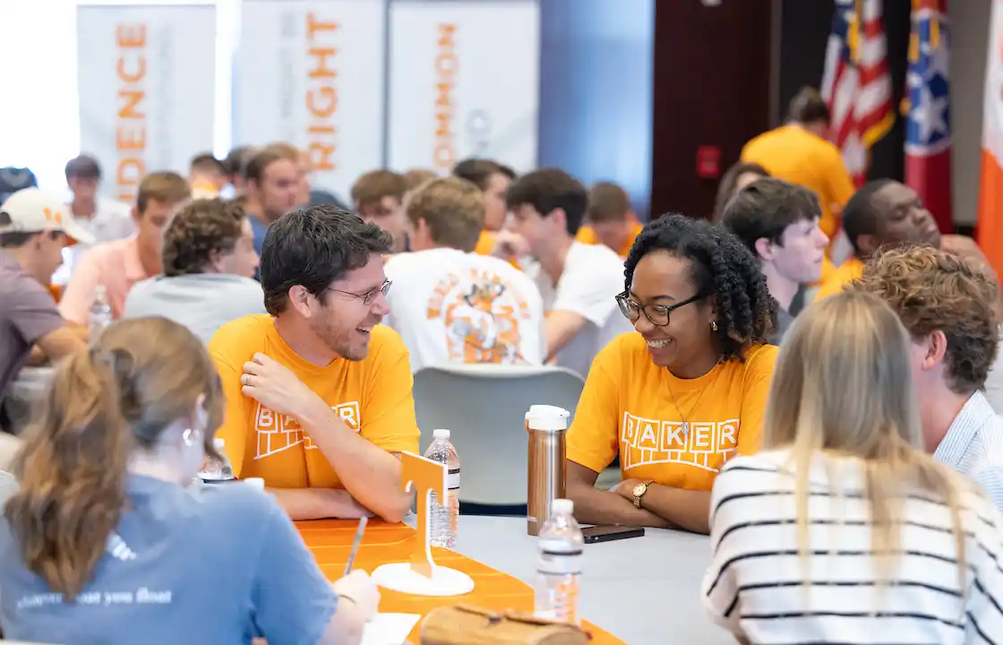 Two professors in focus laughing in UT shirts, students surround them out of focus talking. 