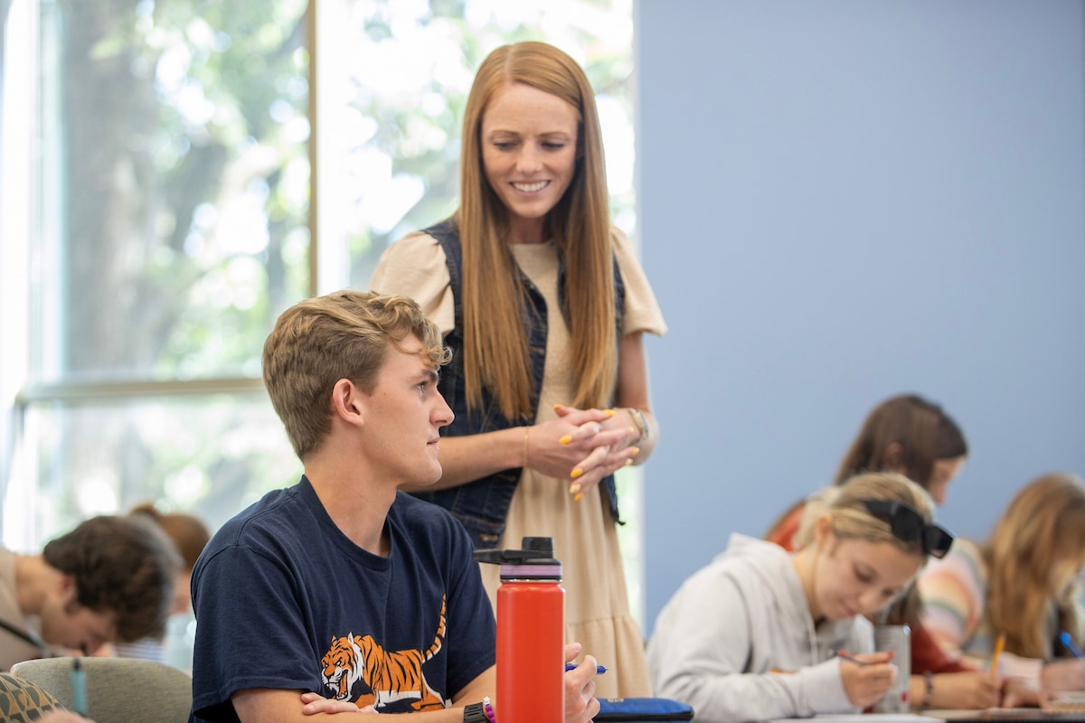 Faculty member standing, speaking to a student sitting down at a desk in the classroom