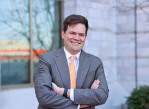 John Stavick poses outside of the Baker School entrance in a gray suit and orange tie.