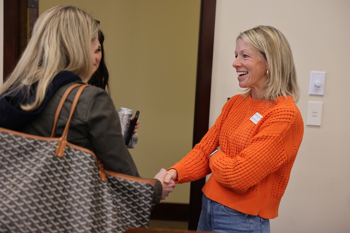 Marianne Wanamaker stands smiling wearing an orange knit sweater shaking a woman's hand. The woman's side faces the camera with a view of her large patterned tote bag. A woman with black hair is slightly visible behind the woman.