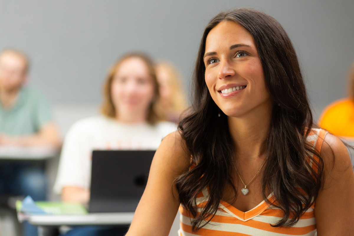 A person with long dark hair smiles while seated in a classroom, with other students blurred in the background.