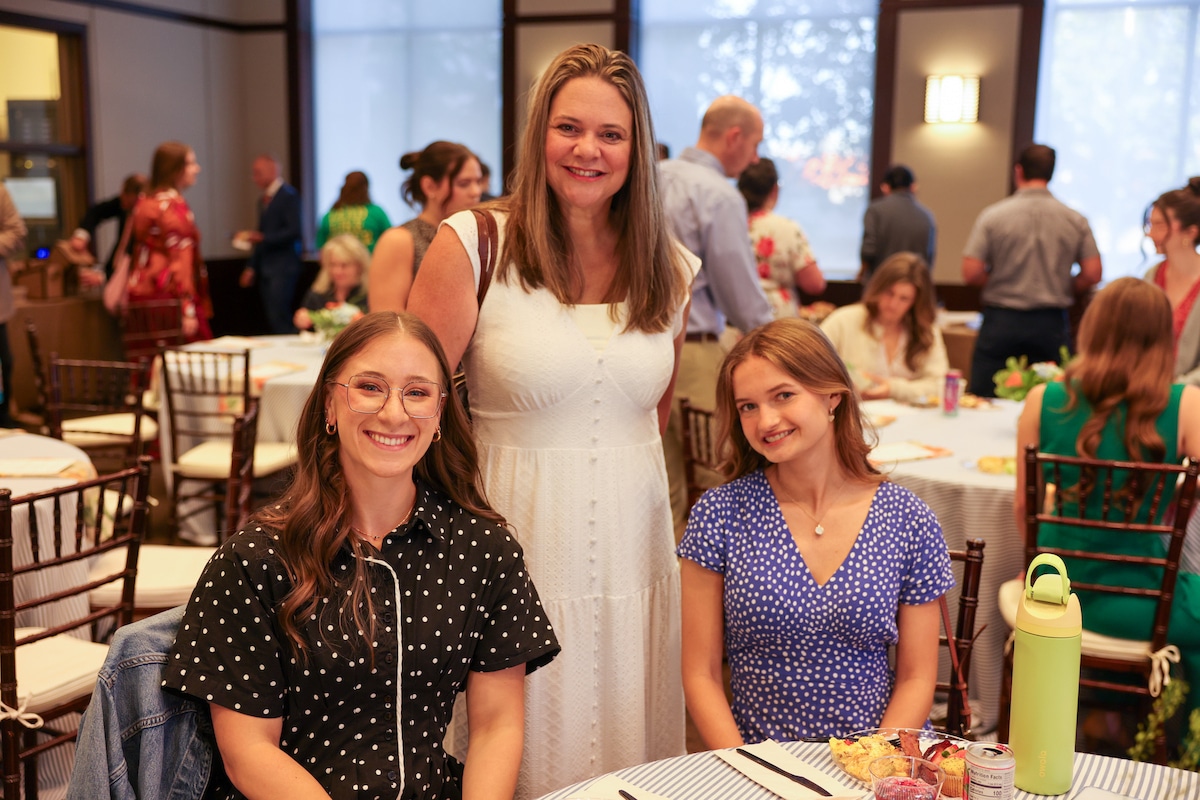 At the Baker Awards, Associate Professor Tara Kolar Bryan stands behind two graduate students. The two students are seated, and everyone is smiling at the camera. Behind them, people sit at tables and go through the buffet line.
