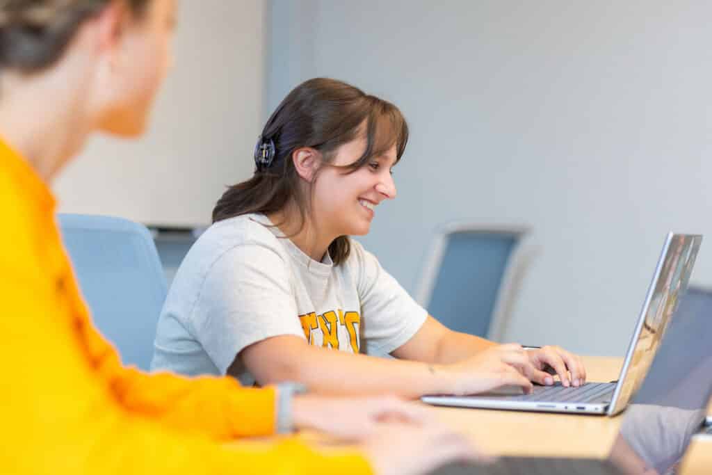 A person with headphones smiles while typing on a laptop in a classroom setting, with another individual seated nearby.