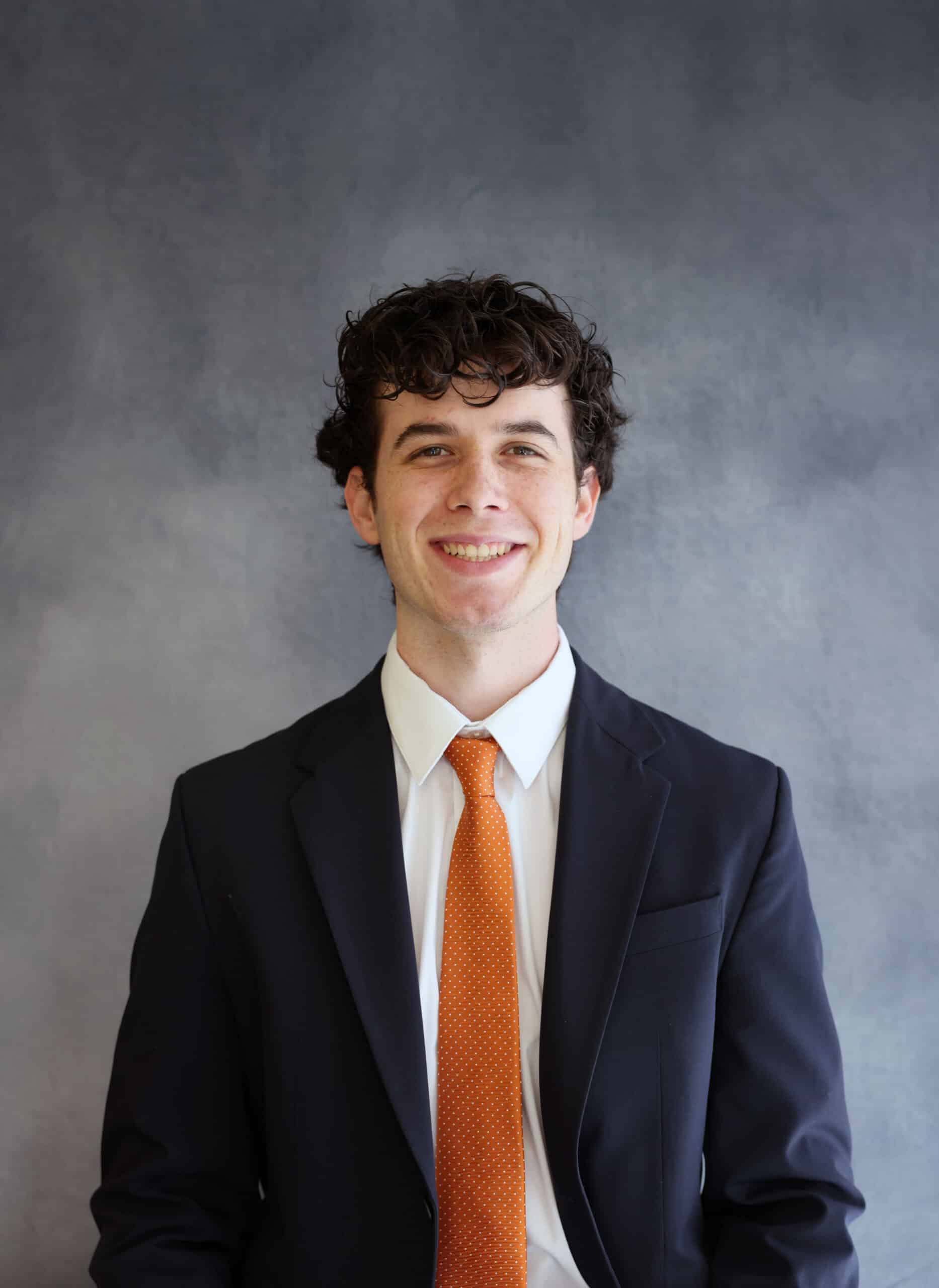 Headshot of Tiago Phielipp. He stands in front of a backdrop, wearing a black suit and an orange tie.